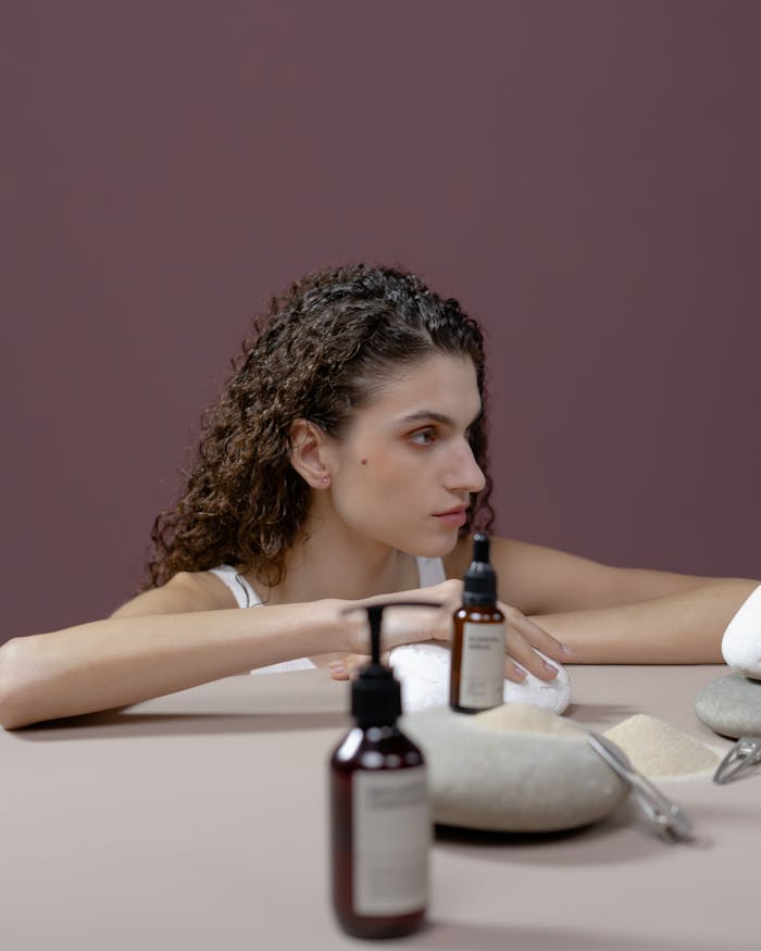 Brunette woman with curly hair posing with cosmetic products, including shampoo, on a minimalist set.